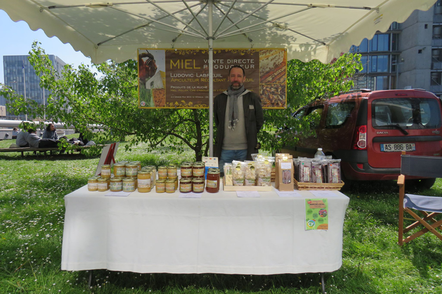 un apiculteur sourit derrière son stand où l'ont peut y voir de nombreuses sortes de miels en bocaux