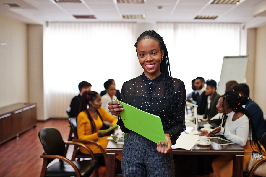 Une jeune femme sourit face à l'objectif. Derrière elle se trouve une réunion avec des personnes assises autour d'une table